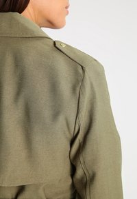 Marynarka z teksturowanej tkaniny w kolorze oliwkowym, z guzikami na naramiennikach i dopasowanym krojem, ukazująca tył oraz część kołnierza.