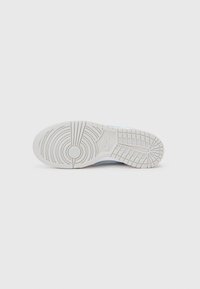 Bílá podrážka tenisky Nike s kulatými a mřížkovými vzory, umístěná na plocho na jednobarevném světlém šedém pozadí.