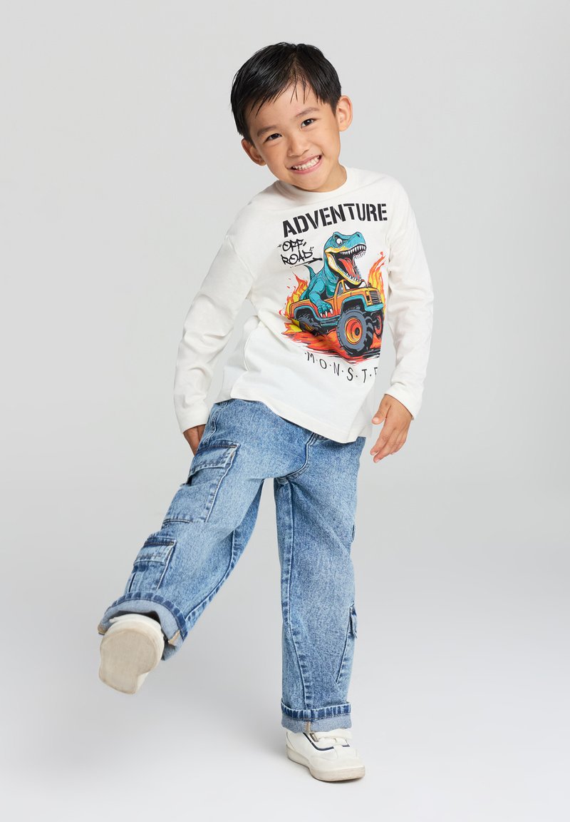 Biała koszula z długim rękawem z grafiką dinozaura i napisem "PRZYGODA", w połączeniu z jasnoniebieskimi dżinsami cargo z kieszeniami i podwiniętymi mankietami.