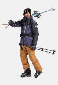 Man wearing ski gear carries skis on shoulder and holds ski poles, dressed in purple jacket, brown pants, gloves, helmet, and ski boots.