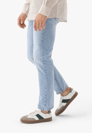 Osoba ubrana w jasnoniebieskie dżinsy i białe trampki z szarymi i ciemnozielonymi akcentami, idąca na tle jednolitego białego tła.