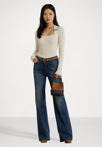 Vrouw in een crèmekleurige ribbeltrui met vierkante halslijn, hoge taille wijdvallende jeans, een bruine riem, bruine schoenen en een bruin en zwarte handtas.