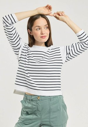 Femme en pantalon vert et chemise blanche à manches longues avec des rayures noires, levant les bras au-dessus de la tête, regardant légèrement sur le côté.