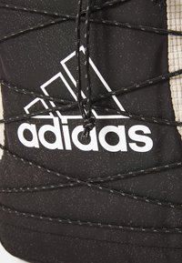Črn športni nahrbtnik z teksturirano površino, ki ima belo logotip Adidas in črne vezalke, tkane čez sprednji del.
