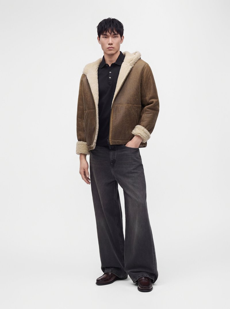 Man wearing brown shearling jacket with rolled cuffs, black polo shirt, wide-leg dark jeans, and dark shoes standing against a plain background.