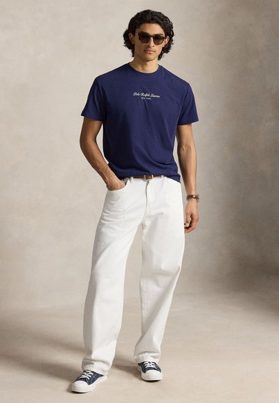 Άνδρας που φοράει σκούρο μπλε μπλουζάκι Polo Ralph Lauren, άσπρο παντελόνι, γυαλιά ηλίου και μπλε αθλητικά παπούτσια, στέκεται με το ένα χέρι στην τσέπη.