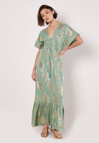 Robe longue vert menthe avec des motifs de feuilles dorées métalliques, décolleté en V profond, manches courtes à volants et ourlet festonné. Tissu léger.