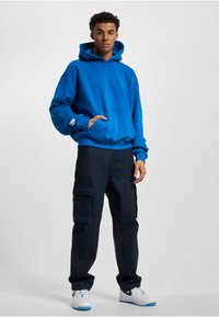 Hanorac albastru cu glugă, cu un buzunar frontal, asortat cu pantaloni cargo negri ce au buzunare laterale, și sneakersi albi cu accente albastre.
