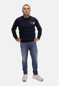 Maglione lavorato a maglia blu navy con ricamo "Il tempo è denaro", abbinato a jeans blu e sneakers grigie. Il look è casual e rilassato.