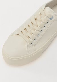 Sneaker en cuir blanc avec une surface texturée, bout rond et lacets assortis. Présente des œillets et une semelle en caoutchouc dans une teinte claire discrète.
