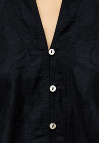 Nahaufnahme eines schwarzen Shirts mit dezenten Blattmustern und drei schimmernden weißen Knöpfen, die über einer V-Ausschnittöffnung geschlossen sind.