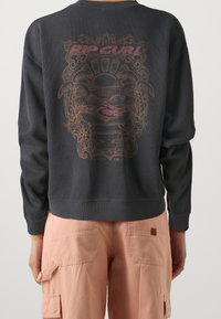 Osoba oblečená v černém topu s dlouhým rukávem, na kterém je vybledlý grafický motiv tropického západu slunce a nápis "Rip Curl" na zadní straně, v kombinaci s světle růžovými cargo kalhotami.