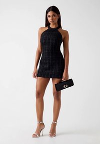 Robe mini noire à col halter avec tissu texturé, coupe ajustée et coutures latérales. Associée à une pochette noire ornée d'un logo doré et des talons argentés.