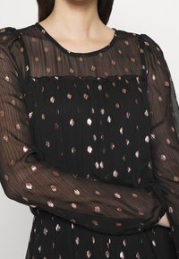 Vestido negro con mangas largas transparentes, con lunares metálicos en oro rosa. Tejido texturizado con detalles fruncidos en el escote.