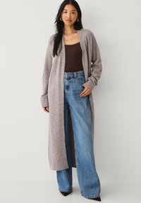 Long, open-front cardigan in light gray knit, featuring wide sleeves and ribbed cuffs, worn over a brown top and high-waisted blue jeans.