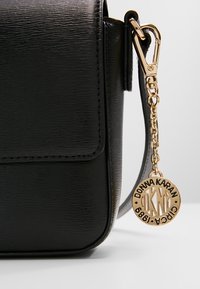 Borsa a tracolla in pelle nera con superficie testurizzata, dotata di un charm dorato con il logo "Donna Karan" e un gancio per il fissaggio.