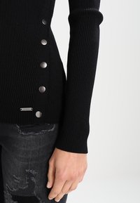 Pull noir côtelé avec des boutons en métal argenté le long de la couture latérale, porté avec un jean gris délavé et usé.