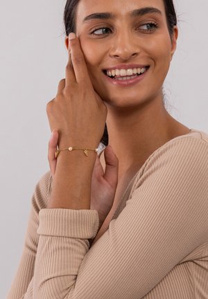 Bracelet en or avec une perle centrale et des accents en forme de feuille, porté au poignet. Le mannequin porte un haut texturé beige clair et sourit.