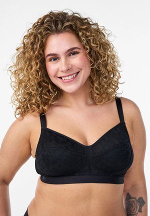 Uśmiechnięta kobieta o kręconych blond włosach, ubrana w czarny koronkowy biustonosz, pokazująca tatuaż róży na lewym przedramieniu na białym tle.