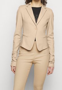 Mujer vestida con una chaqueta beige ajustada y pantalones a juego sobre una camiseta negra con texto dorado, de pie frente a un fondo claro y liso.