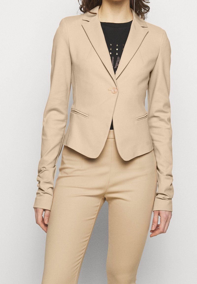 Mujer vestida con una chaqueta beige ajustada y pantalones a juego sobre una camiseta negra con texto dorado, de pie frente a un fondo claro y liso.