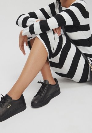 Personne assise portant une robe à rayures noires et blanches et des baskets noires avec les initiales "RKO" sur le côté.