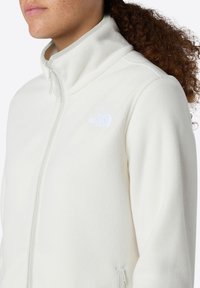 Jachetă din fleece alb cu guler înalt și fermoar complet. Prezintă un mic logo alb pe piept și un material moale, texturat.
