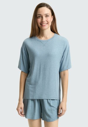 Lachende vrouw met lang bruin haar, gekleed in een lichtblauw kort mouw T-shirt en bijpassende short, staand tegen een lichte achtergrond.