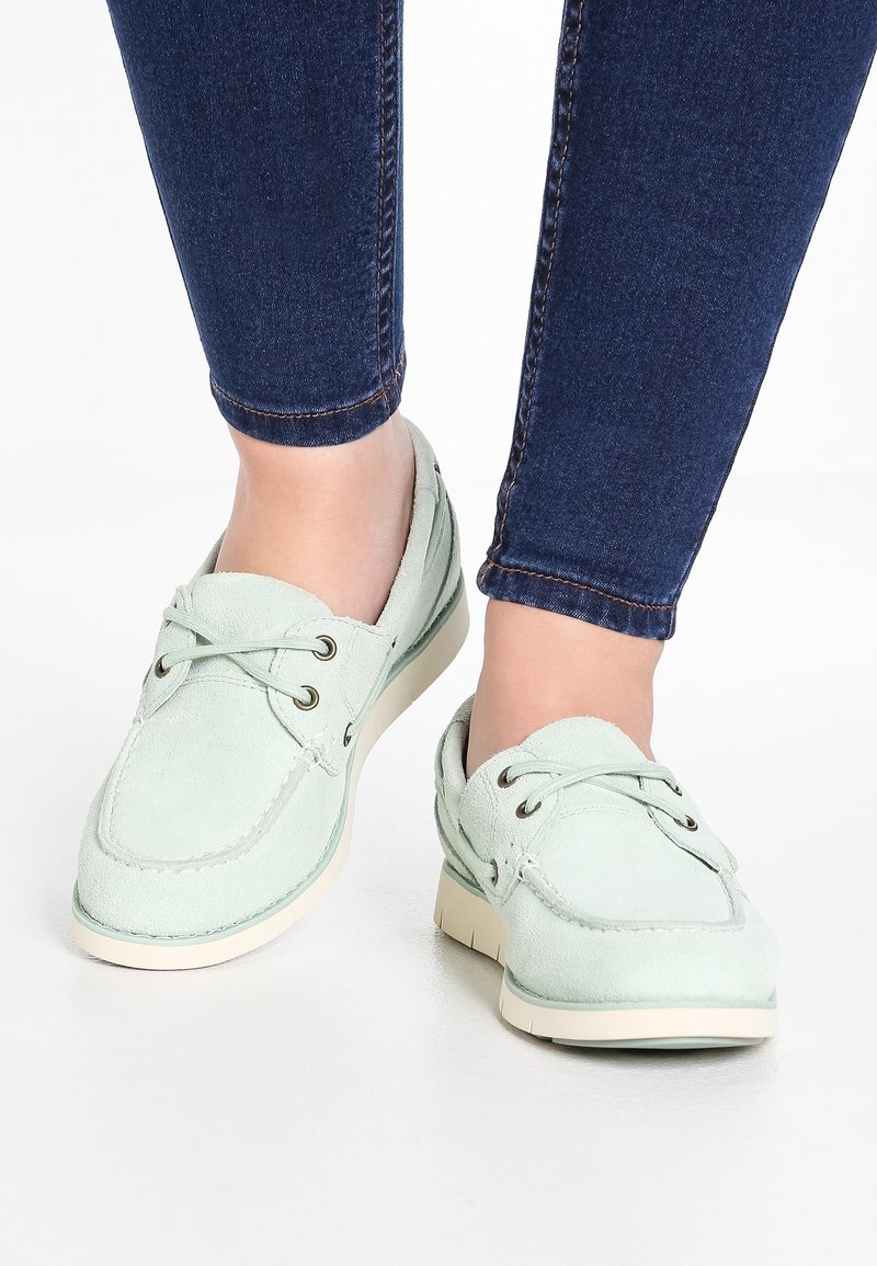 Pieds portant des mocassins en daim vert clair associés à un jean skinny en denim bleu foncé sur un fond blanc.