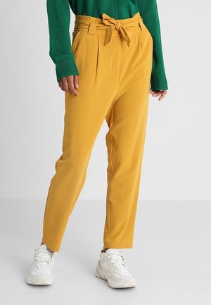 Personne portant un pantalon taille haute jaune moutarde avec une ceinture nouée, un pull vert à manches longues et des baskets blanches épaisses, debout sur un sol gris.