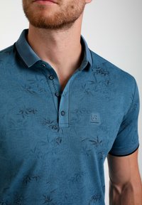 Polo bleu avec un motif de feuilles subtil, fabriqué en tissu doux. Il présente un col, trois boutons et un logo sur la poitrine.
