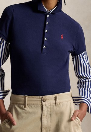 Persoon die gelaagde kleding draagt met een marineblauw poloshirt over een blauw-wit gestreept shirt met lange mouwen en beige broek, handen in de zakken.