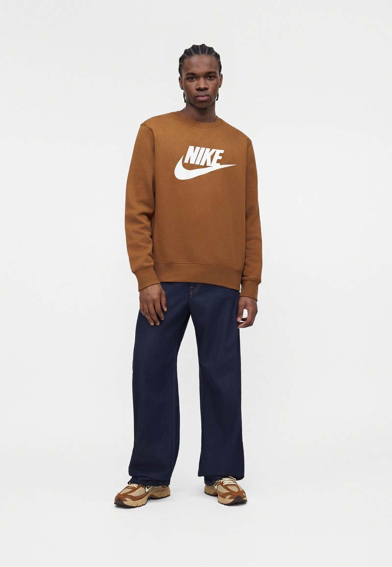 Junger Mann steht und trägt einen braunen Nike-Sweatshirt, dunkelweite Jeans und beige, braune und weiße Sneaker vor weißem Hintergrund.