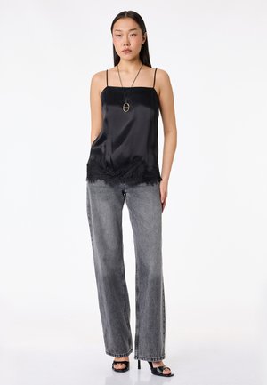 Młoda kobieta stoi, ubrana w czarną satynową koszulkę na ramiączkach z koronkową lamówką, szare szerokie jeansy oraz czarne sandały na obcasie i naszyjnik z zawieszką.