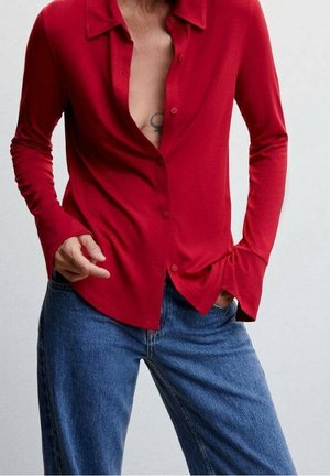 Osoba nosící červenou košili s dlouhými rukávy rozepnutou částečně až na tělo a modré džíny, s jednou rukou v kapse a viditelným tetováním na hrudi.