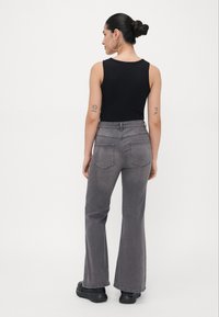 Jean taille haute greige avec jambes évasées, design à cinq poches, coutures tonales et texture usée, associé à un débardeur noir ajusté.