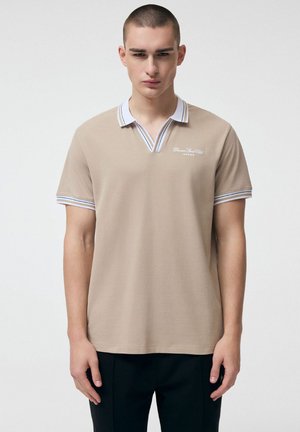 Młody mężczyzna o krótkich włosach, ubrany w beżową koszulkę polo z białym kołnierzykiem i mankietami z niebieskimi paskami, stoi na jednolitym tle.