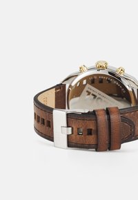 Bracelet de montre en cuir marron avec des découpes, boucle en métal argenté et boutons chronographes dorés ; boîtier en acier inoxydable avec fond transparent.