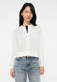 Cardigan léger blanc avec des manches longues et un col rond, doté de boutons sur le devant et d'un petit logo sur la poitrine.