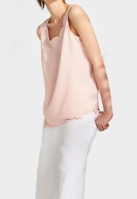 Person som bär en ärmlös ljusrosa topp med scalloped-kant och lösa vita byxor mot en enfärgad ljus bakgrund.