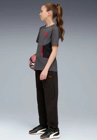 Grijze sportshirt met zwarte accenten en een rode ritszak, gecombineerd met zwarte joggingbroeken met zijzakken en een rood logo, terwijl hij een voetbal vasthoudt.