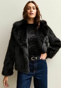 Svart fuskpälsjacka med en bred krage, texturerad yta och långa ärmar, i kombination med en mönstrad topp och högmidjade jeans.