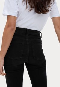 Højtaljede sorte denimjeans med en tætsiddende pasform, som har to baglommer og en glat tekstur. En hvid t-shirt er delvist synlig.
