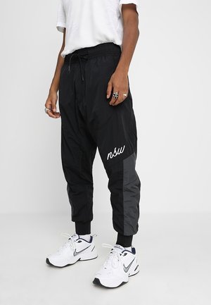 Persona con pantalones negros y grises con cordón y texto "nsw", y zapatillas blancas de Nike, de pie sobre un suelo blanco contra un fondo blanco.