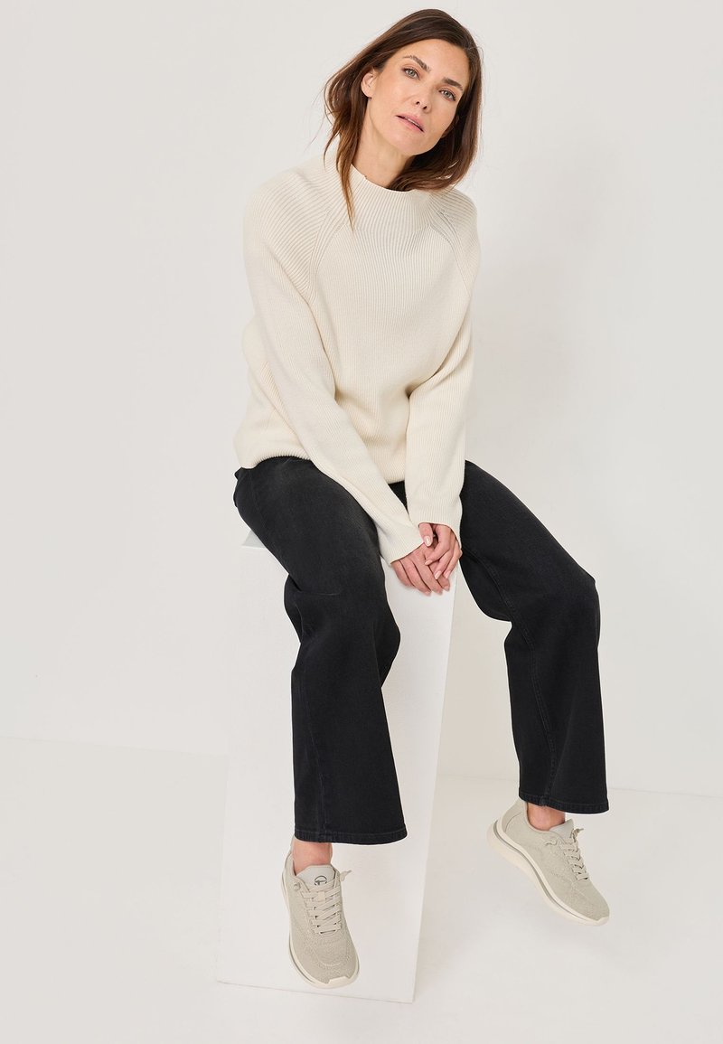 Krēmveida ribota džemperis, melnas platas bikses un gaiši pelēki čības. Sēdoša poza uz baltas pjedestāla, neitrāls fons.