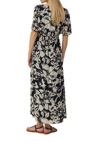 Frau mit langen blonden Haaren, die ein schwarz-weißes Blumenkleid im Maxistil und schwarze flache Sandalen trägt, steht mit dem Rücken nach vorne.
