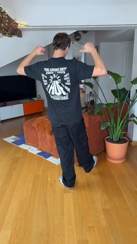 Black t-shirt with white graphic print on the back; loose black pants; light-colored sneakers. Indoor setting with plants and furniture.