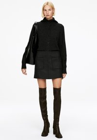 Dunkelstrickjacke mit hohem Kragen, Knopfverschluss und übergroßen Ärmeln. Kombiniert mit einem figurbetonten, anthrazitfarbenen Rock und knielangen braunen Stiefeln.