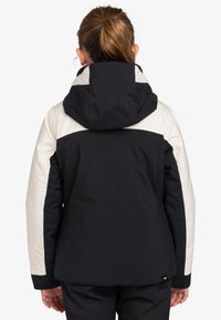 Veste isolante noire et crème avec capuche, présentant un design en blocs de couleurs, un tissu texturé et un accent logo à l'ourlet.