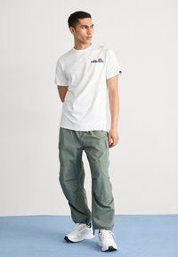 T-shirt bianca in cotone con logo "ellesse" blu, abbinata a pantaloni cargo verde oliva con tasche e polsini elasticizzati, completata da sneakers bianche.